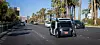 Zoox autonomous shuttle driving on a sunny multi-lane city street with palm trees.