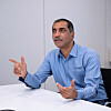 Man in blue shirt gesturing as he speaks at a white table in a plain office room