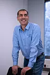 Man in blue shirt stands by a chair in a bright office meeting room.