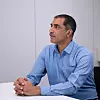 Man in blue branded shirt sits at white table in plain office setting