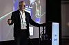 Man in suit speaking on stage next to a lit lectern and large presentation screen showing Jim Foresi
