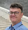 Man in glasses stands near stone steps with a modern wall behind him