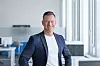 Smiling man in a blazer stands in a bright open-plan office near large windows.