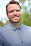 Smiling man in a blue shirt standing outside with blurred greenery behind him