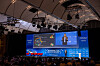 Wide view of a conference stage with a presenter and large blue presentation screen in an auditorium.