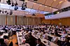 Crowded conference hall with seated attendees at long tables under overhead lighting.