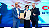 Two presenters hold a framed certificate on stage in front of a large branded event screen.
