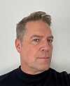 Close-up portrait of a man in a black top against a plain white wall.