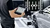Technician holding a tablet next to a grey car in a workshop with diagnostic lights.