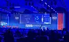 Blue-lit conference stage with AEK screens and audience seated below.