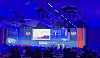 Blue-lit conference hall with a large stage screen, branded panels and an audience in silhouette.