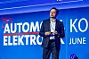 A man in a suit stands on stage in front of a blue automotive electronics event backdrop.
