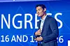 Man in a suit speaking on stage before a blue conference backdrop.