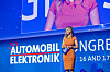 A woman in an orange dress stands on stage beneath a large Automobil-Elektronik Congress screen.