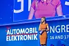 A woman in an orange dress stands on stage beneath a large Automobil-Elektronik Congress screen.