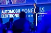 Presenter on a blue-lit stage at Automobilkongress Elektronik with audience silhouettes in front.