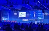 Conference audience facing a blue-lit stage with presentation screens and a speaker.