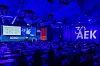 Audience seated in a conference hall facing a stage with large illuminated screens and blue lighting.
