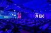 Large audience seated at tables in an indoor conference hall with a lit stage and presentation screens.