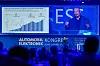 Speaker on stage beside a chart and conference branding during a blue-lit business presentation.