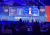 Conference stage with blue lighting, large screens, AEK branding and seated audience.