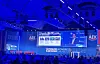 Blue-lit conference stage with large screens, AEK branding and an audience seated below.