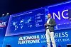 Speaker on stage beside a large screen at an automotive electronics congress.