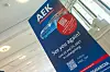 Tall blue AEK exhibition banner in a bright indoor space with stairs and windows.