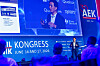 Speaker onstage at a congress with a large blue presentation screen and audience silhouettes in the foreground.