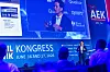 Speaker onstage at a congress with a large blue presentation screen and audience silhouettes in the foreground.