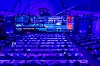 Wide view of a conference hall with rows of tables, a lit stage and large screens.