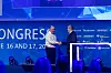 Two men shake hands on a conference stage in front of a blue backdrop with sponsor logos.