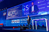 Presenter on a blue-lit conference stage with large screens and sponsor logos.