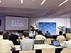 Conference audience seated in a meeting room watching a presentation beside an ALC-branded screen.