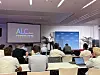 Conference audience seated in a meeting room watching a presentation beside an ALC-branded screen.
