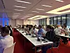 People seated at tables in a conference room with blue lighting and large windows.