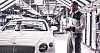 Worker holding a clipboard beside a white luxury car in a factory.