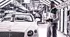 Worker holding a clipboard beside a white luxury car in a factory.