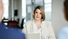 Woman in a white blazer seated at a meeting table with blurred people in the foreground.