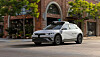 White self-driving test car moving along a city street beside brick buildings and trees.