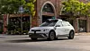 White self-driving test car moving along a city street beside brick buildings and trees.