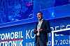 A man stands on stage holding a small device in front of a blue conference backdrop.
