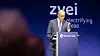 A man in a suit speaks at a podium on a stage with a blue backdrop and the words ‘zwei’ and ‘electrifying ideas’.