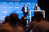 Two men speaking on a conference stage before a blue logo backdrop, with audience silhouettes in front.