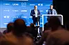 Two men speaking on a conference stage before a blue logo backdrop, with audience silhouettes in front.