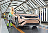 Pink electric car on an indoor factory line with workers nearby and structural lighting overhead.