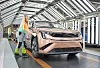 Pink electric car on an indoor factory line with workers nearby and structural lighting overhead.