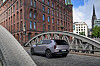 Grey SUV driving on a cobblestone bridge with a red-brick building and city skyline in the background.