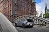 Grey SUV driving on a cobblestone bridge with a red-brick building and city skyline in the background.