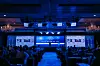 Conference audience facing a stage with large screens and blue lighting in a ballroom.